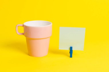 Somon bardağı ve mavi bir çamaşır iğnesi içinde boş bir not kağıdı. Kare dikdörtgen şeklinde renkli anımsatıcı sarı bir arka plan. Boş alan ve ofis malzemeleri.