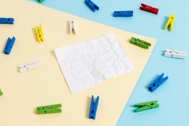 Mavi arka plan ve sarı kare ortasında boş dikdörtgen şeklinde hatırlatma notu etrafında yerleştirilen renkli clothespin. Ofis malzemeleri, bir bağlantı elemanı tarafından sabitlenmiş boş kağıt.