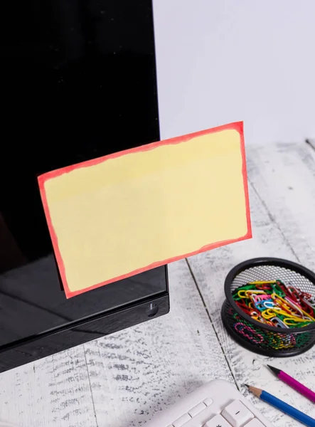 표기 용지는 흰색 키보드 근처의 검은 색 컴퓨터 화면에 붙어 있으며 나무 테이블 위에 고정되어 있습니다. PC 모니터는 그것에 노란색 노트 시트 테이프와 쓰기 장비 근처에 배치. — 스톡 사진
