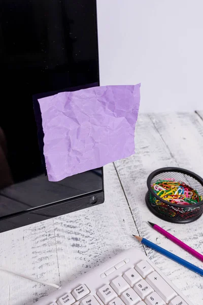 Notatie papier stick naar zwart computerscherm in de buurt van witte toetsenbord en stationair boven de houten tafel. PC-monitor geplaatst in de buurt van schrijf apparatuur met paarse notitie blad tape erop. — Stockfoto