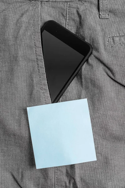 Mavi bir not kağıt yakınında resmi çalışma pantolon ön cebinde siyah akıllı telefon cihazı. Modern gadget notasyon levha yakın erkek pantolon içine yerleştirilir — Stok fotoğraf