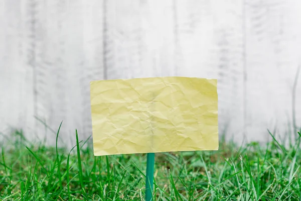 Plain paper cardboard attached to a stick and plugged in the grassy land. Yellow empty sheet is placed in ground with green grass. Photography idea with small object