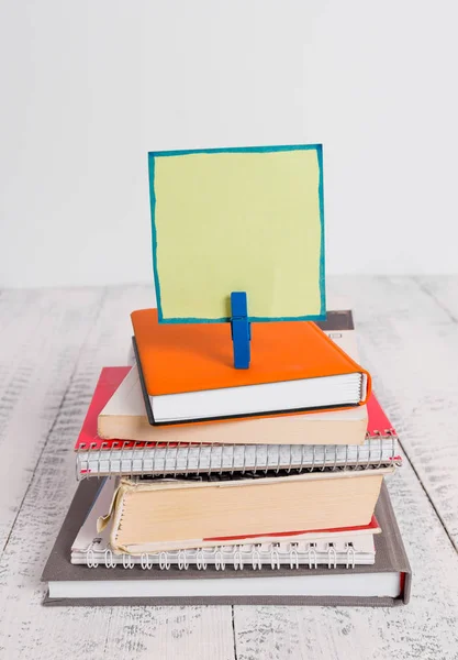 Square shaped paper above a pile of notebooks fixed by a blue clothespin. Different books stacked under an empty reminder colorful note. White wooden floor and white background.