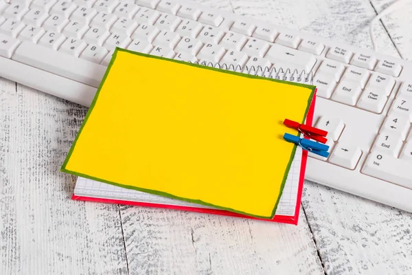 Cuadrado cuaderno de papel con un rectángulo en forma de nota vacía por encima fijado por dos pinzas de ropa en la hoja. Teclado blanco bajo un copybook y fondo de madera claro . —  Fotos de Stock