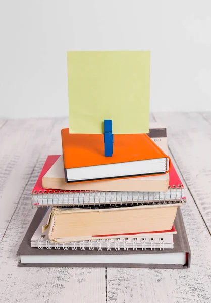Square shaped paper above a pile of notebooks fixed by a blue clothespin. Different books stacked under an empty reminder colorful note. White wooden floor and white background.