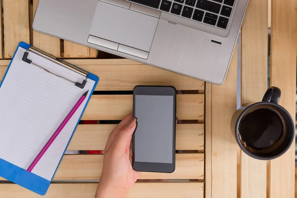 Ung dam använder och håller en mörk smartphone i en låda bord med en mugg svart kaffe. Kontorsmateriel, mobiltelefon, tekniska enheter och trä skrivbord. — Stockfoto