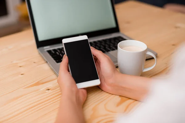 Ung dam håller en smartphone med båda händerna. Grå bärbar dator på bakgrunden med svart tangentbord och en mugg kaffe. Kontorsmateriel, tekniska apparater och trä skrivbord. — Stockfoto
