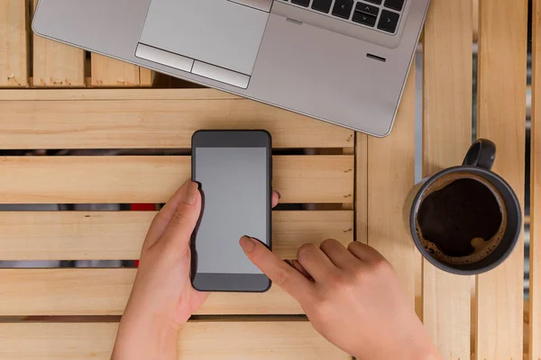 Ung dam använder och håller en mörk smartphone i en låda bord med en mugg svart kaffe. Kontorsmateriel, mobiltelefon, tekniska enheter och trä skrivbord. — Stockfoto