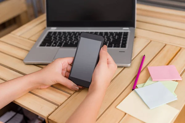 Ung dam använder och håller en mörk smartphone i en låda bord med en mugg svart kaffe. Kontorsmateriel, mobiltelefon, tekniska enheter och trä skrivbord. — Stockfoto