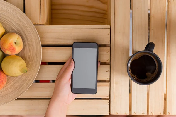 Ung dam använder och håller en mörk smartphone i en låda bord med en mugg svart kaffe. Kontorsmateriel, mobiltelefon, tekniska enheter och trä skrivbord. — Stockfoto