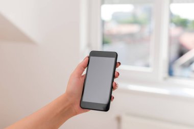 Genç bayan evin içinde karanlık bir akıllı telefon kullanıyor. Evin ambiyansında siyah bir cep telefonu tutan kadın. Boş alan ile teknolojik cihazlar ve ofis malzemeleri ile Kız.