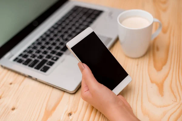 Jeune femme tenant un smartphone avec la main gauche. Ordinateur portable gris sur le fond avec clavier noir, une tasse de café, fournitures de bureau, appareils technologiques et bureau en bois . — Photo