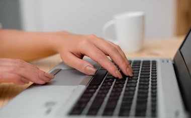 Genç bayan gri bir dizüstü bilgisayar kullanarak ve arka planda kahve veya çay bir fincan ile siyah klavye yazarak. Ofis malzemeleri, teknolojik cihazlar ve ahşap masa.