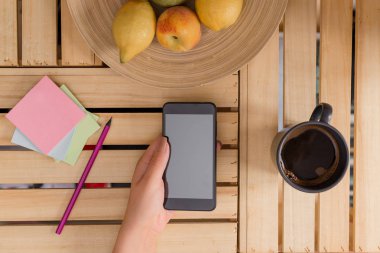 Genç bayan kullanarak ve siyah kahve bir fincan ile bir sandık masada karanlık bir akıllı telefon tutarak. Ofis malzemeleri, cep telefonu, teknolojik cihazlar ve ahşap masa.