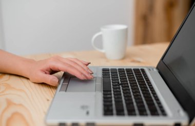 Dokunmatik yüzeyde eliyle gri bir dizüstü bilgisayar ve siyah klavye kullanan genç bayan. Arka planda bir fincan kahve. Ofis malzemeleri, teknolojik cihazlar ve ahşap masa.