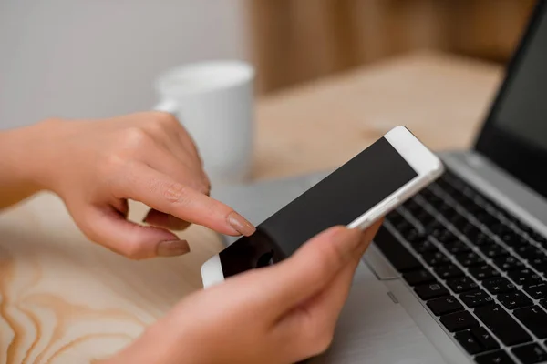 Jovencita usando un smartphone y tocando la pantalla. Ordenador portátil gris en el fondo con teclado negro y una taza de café. Materiales de oficina, dispositivos tecnológicos y escritorio de madera . — Foto de Stock