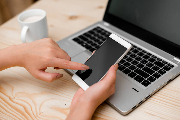 Young lady using a smartphone and touching the screen. Gray laptop computer on the background with black keyboard and a mug of coffee. Office supplies, technological devices and wooden desk.