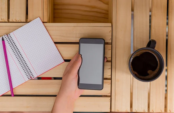 Νεαρή κυρία χρησιμοποιώντας και κρατώντας ένα σκοτεινό smartphone σε ένα κιβώτιο τραπέζι με μια κούπα του μαύρου καφέ. Αναλώσιμα γραφείου, κινητό τηλέφωνο, τεχνολογικές συσκευές και ξύλινο γραφείο. — Φωτογραφία Αρχείου