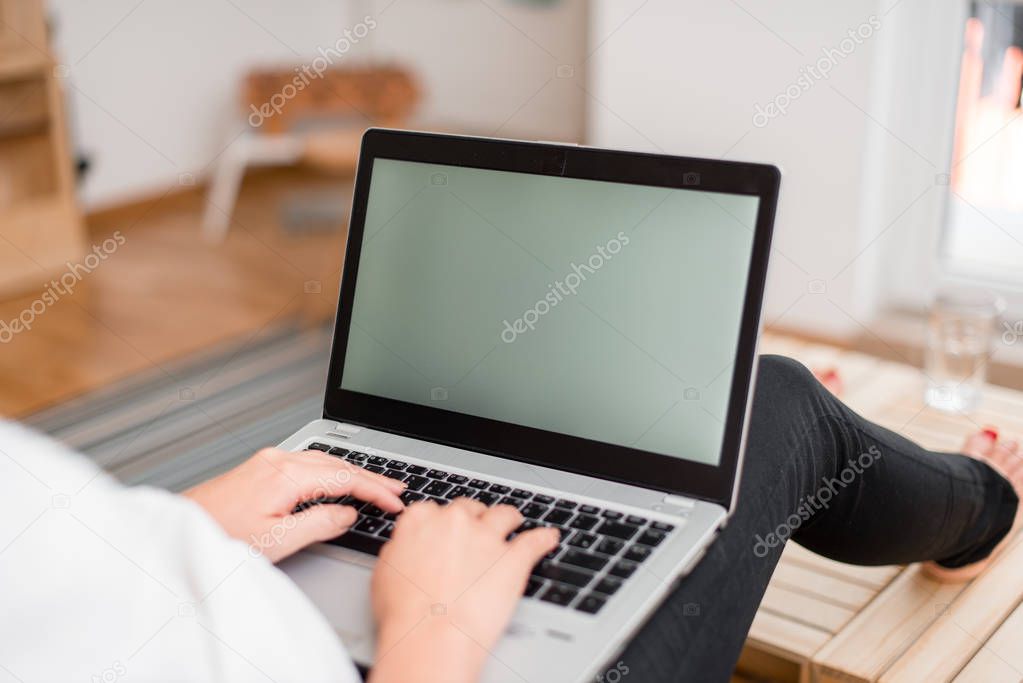 Young lady using a gray laptop computer and typing in the black keyboard with both hands in a room. Woman in a house ambient with office supplies and technological devices.