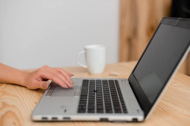 Dokunmatik yüzeyde eliyle gri bir dizüstü bilgisayar ve siyah klavye kullanan genç bayan. Arka planda bir fincan kahve. Ofis malzemeleri, teknolojik cihazlar ve ahşap masa.