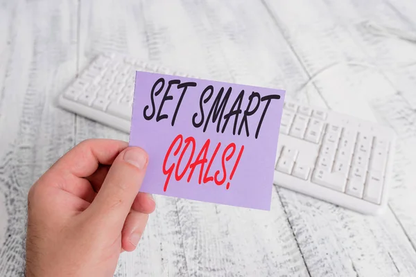 Text sign showing Set Smart Goals. Conceptual photo list to clarify your ideas focus efforts use time wisely man holding colorful reminder square shaped paper white keyboard wood floor.