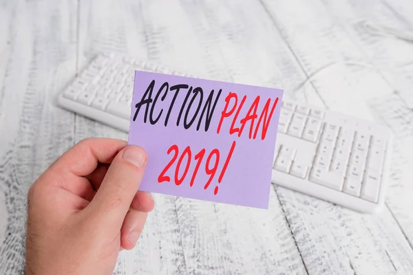 Text sign showing Action Plan 2019. Conceptual photo proposed strategy or course of actions for current year man holding colorful reminder square shaped paper white keyboard wood floor.