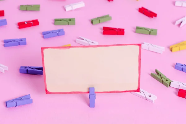 Farbige Wäscheklammer um das leere rechteckige quadratische Erinnerungsstück in der Mitte eines rosafarbenen Hintergrundes platziert. Bürobedarf, unbedrucktes Papier, das mit einem Verschluss befestigt ist. — Stockfoto