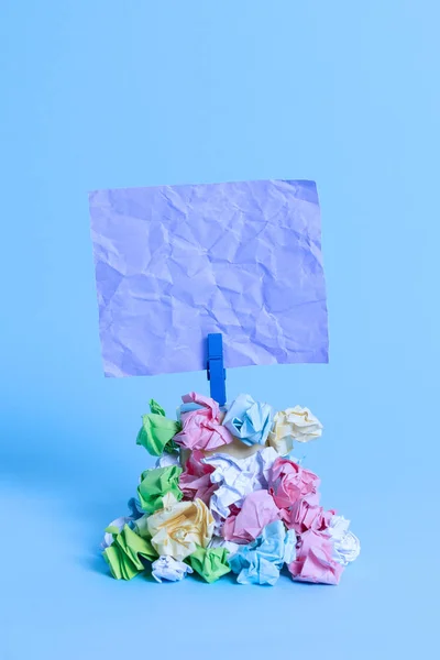 Vierkante vormige herinnering boven een stapel verfrommeld papier, bevestigd door een blauwe wasknijper. Verschillende bladen gestapeld onder een lege kleurrijke noot. Licht blauwe achtergrond. — Stockfoto