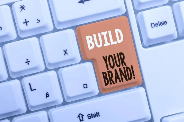 Text sign showing Build Your Brand. Conceptual photo creates or improves customers knowledge and opinions of product White pc keyboard with empty note paper above white background key copy space.