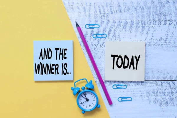 Word writing text And The Winner Is. Business concept for announcing a demonstrating or thing that wins something Notepads marker pen colored paper sheet alarm clock wooden background. — Stock Photo, Image