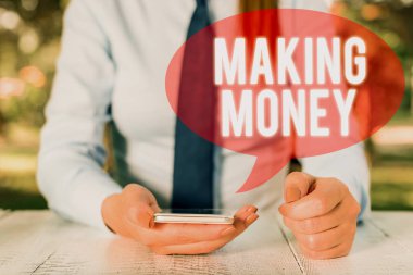 Writing note showing Making Money. Business photo showcasing Giving the opportunity to make a profit Earn financial support Female business person sitting by table and holding mobile phone.
