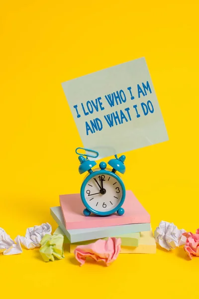 Text sign showing I Love Who I Am And What I Do. Conceptual photo High selfstem being comfortable with your job Alarm clock sticky note paper balls stacked notepads colored background.