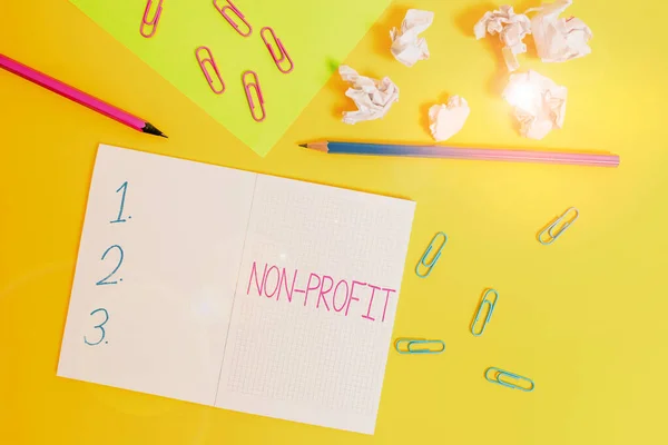 Word writing text Non Profit. Business concept for type of organization that does not earn profits for its owners Blank squared notebook pencils paper sheet crushed balls colored background.