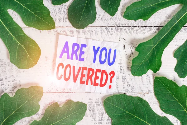 Text sign showing Are You Covered Question. Conceptual photo asking showing if they had insurance in work or life Leaves surrounding notepaper above a classic wooden table as the background.