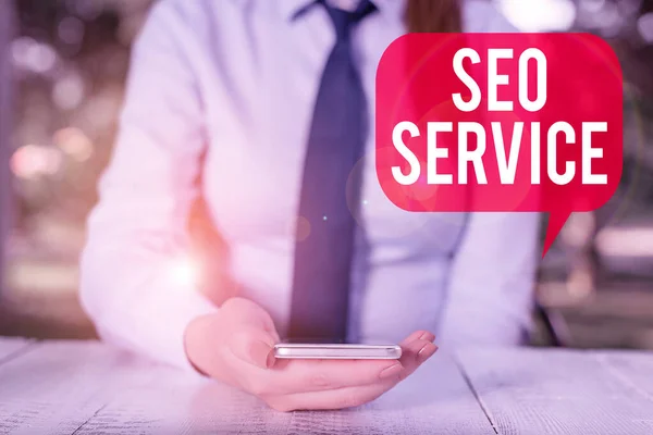 Nota escrita que muestra Seo Service. Foto de negocios mostrando técnicas y procedimientos para aumentar la visibilidad del sitio web Mujer de negocios sentada junto a la mesa y sosteniendo el teléfono móvil . —  Fotos de Stock