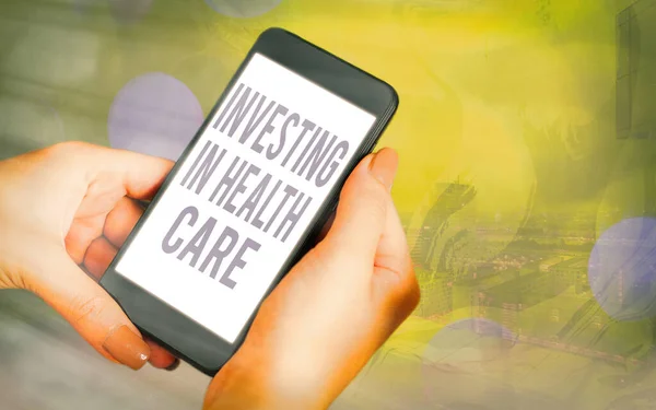 Handwriting text Investing In Health Care. Concept meaning put money on maintenance or improvement of health Modern gadgets with white display screen under colorful bokeh background.