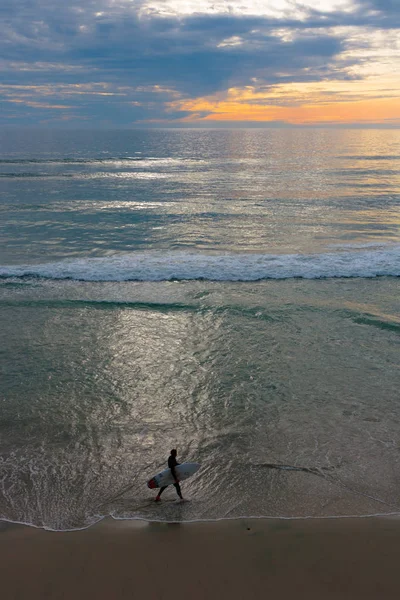 Sunset seascape, orange, blue, yellow sky, with aqua marine green sea, white waves rolling in, surfer at dusk walking in the surf, carrying his white and red surf board