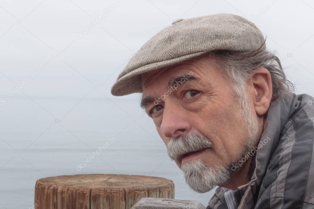 Close up of thoughtful, serious senior man with a gray beard wearing a tweed cap looking at camera with ocean horizon in background