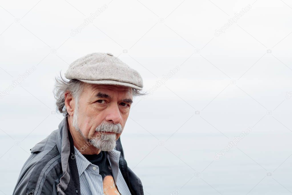 Close up of thoughtful, serious senior man with a gray beard wearing a tweed cap looking at camera, faint ocean horizon in background
