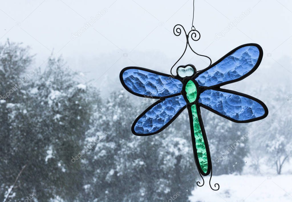 Cold snowy day with oak trees seen through a window with bright blue and green stained-glass dragonfly sun catcher hanging in window