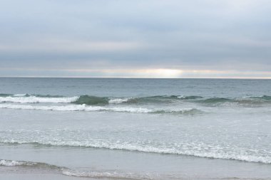 Dalga kıyıya doğru haddeleme, yumuşak ışık mavi bulutlar, ufukta güneş şişer, huzurlu nazik.