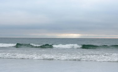 Dalga kıyıya doğru haddeleme, yumuşak ışık mavi bulutlar, ufukta güneş şişer, huzurlu nazik.