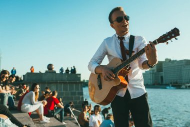 Moskova, Rusya - 9 Mayıs 2018: güneş gözlüğü, kravat ve St. George kurdele ile genç adam karşı riverside gitar çalıyor. Oturma ve onun arkasında oturan insan kalabalığı. Gorki Park İlkbahar geç. Gün