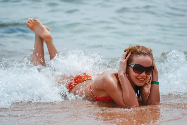 Rire et fille heureuse dans un maillot de bain orange est couché sur la plage de la mer humide et profiter des vagues laver les corps et le soleil d'été lumineux — Photo