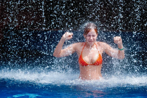 Laughing and happy girl in an orange swimsuit plays jumping and having fun under a waterfall in the pool — Stock Photo, Image