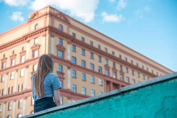 Chica de pelo largo en una camiseta gris y falda negra está sentado en el parapeto de piedra en el fondo de un enorme edificio de varios pisos — Foto de Stock