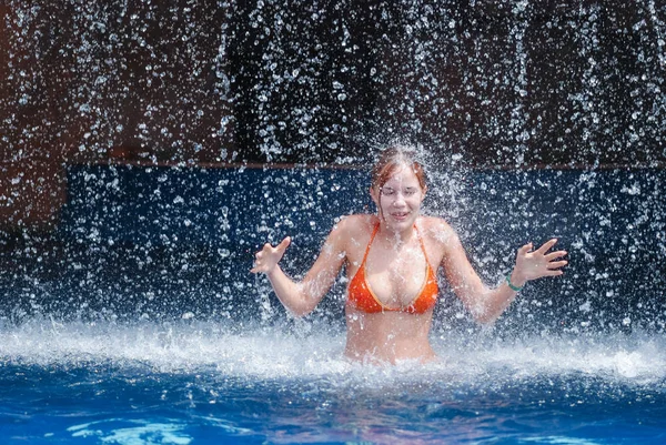 Lachendes und glückliches Mädchen in orangefarbenem Badeanzug spielt Springen und Spaß unter einem Wasserfall im Pool — Stockfoto