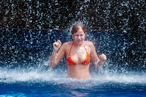 Laughing and happy girl in an orange swimsuit plays jumping and having fun under a waterfall in the pool — Stock Photo, Image