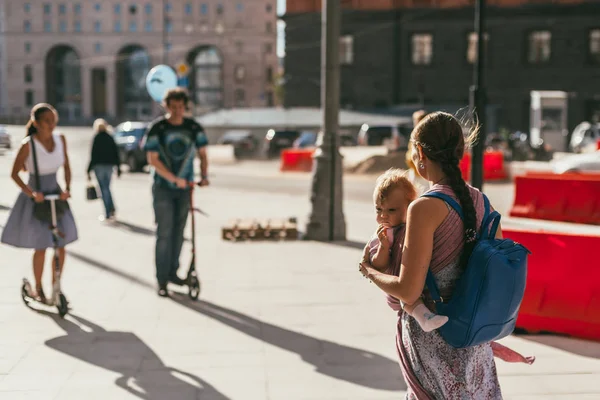 MOSCÚ, RUSIA - 14 de agosto de 2015. Joven madre con un bebé en un cabestrillo en la parte delantera y una mochila a sus espaldas caminando por la ciudad. Un niño y una niña están montando scooters en el fondo — Foto de Stock