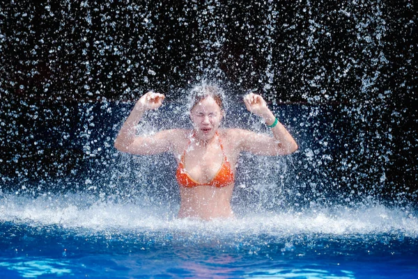 Lachendes und glückliches Mädchen in orangefarbenem Badeanzug spielt Springen und Spaß unter einem Wasserfall im Pool — Stockfoto